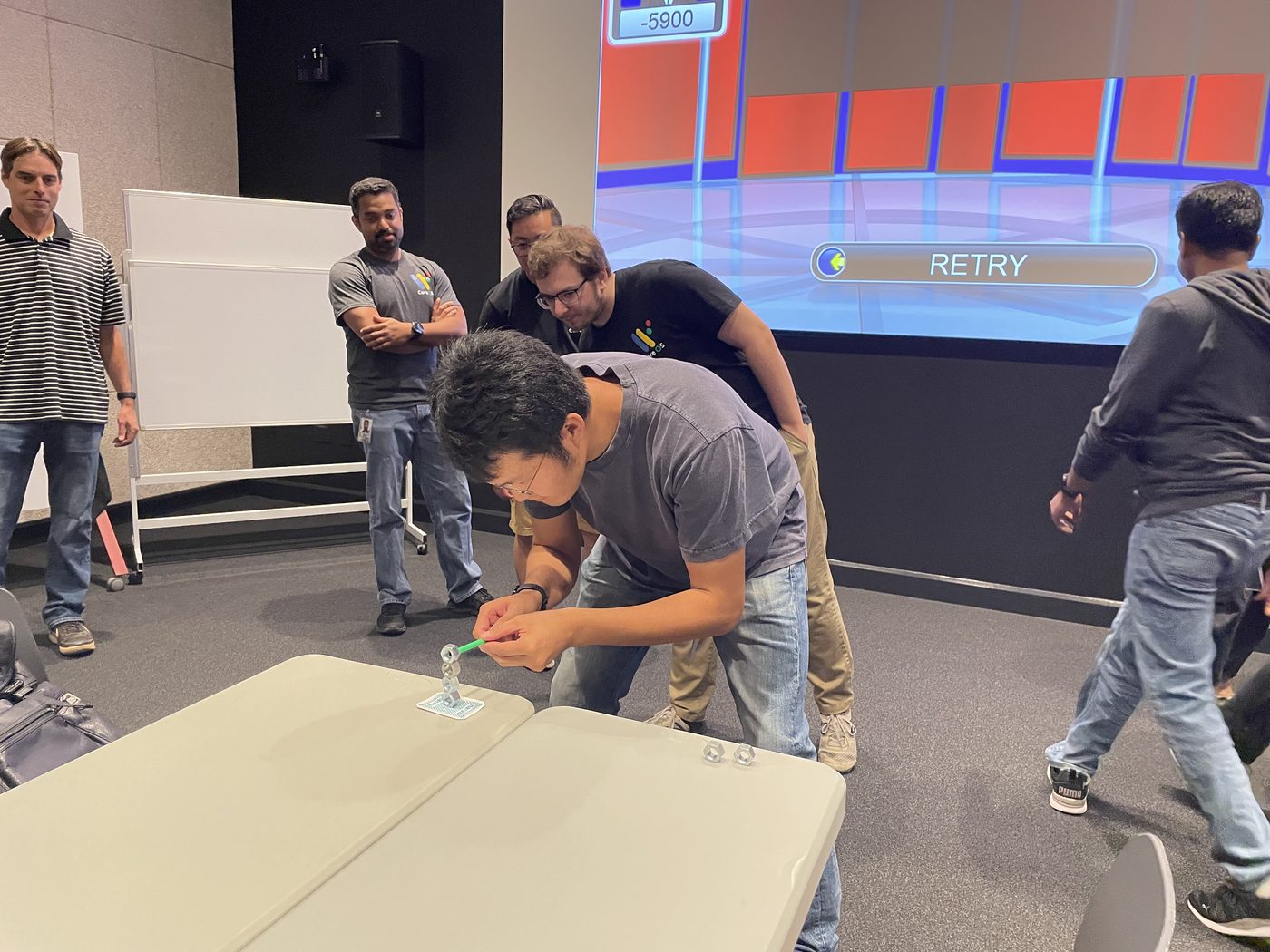 A participant stacks metal pieces in a fast tabletop challenge while teammates watch beside the game board